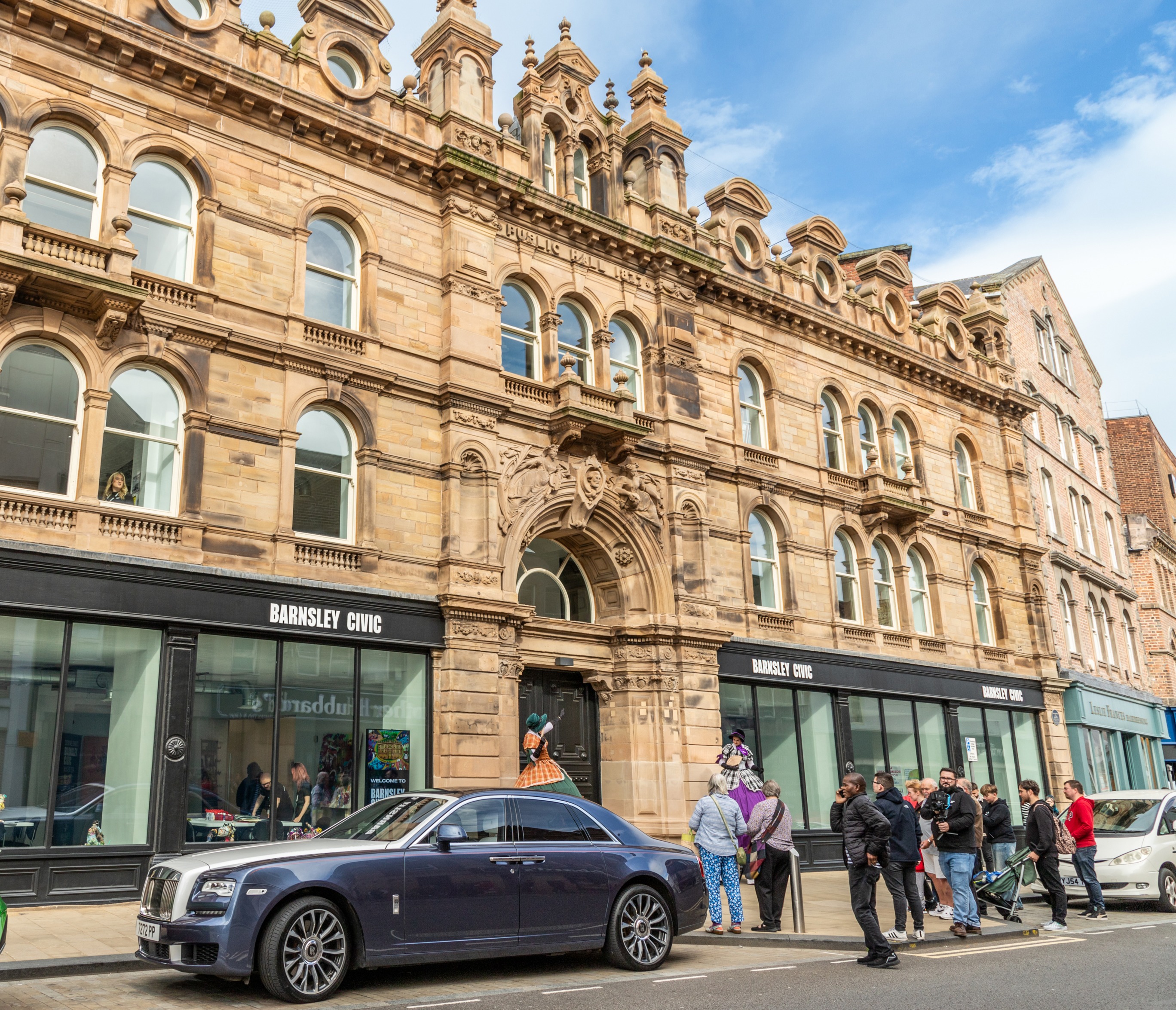 A photograph of the exterior of Barnsley Civic. It's a four-storey Victorian brick building, with a modern glass-front on the ground floor. People are standing on the street watching two performers on stilts.