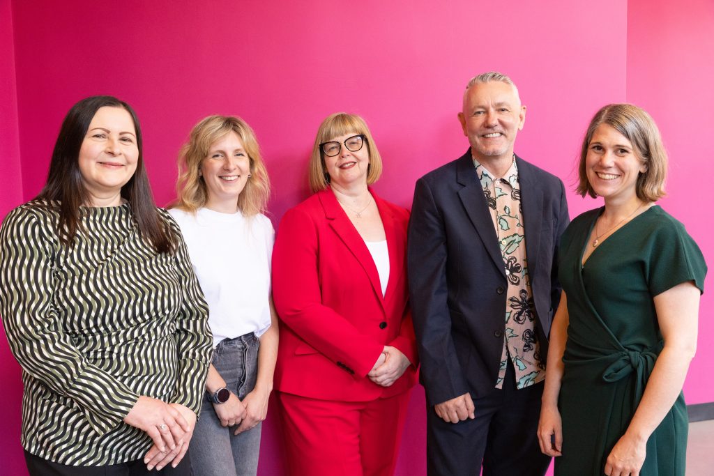 The team smiling at camera in front of a pink background