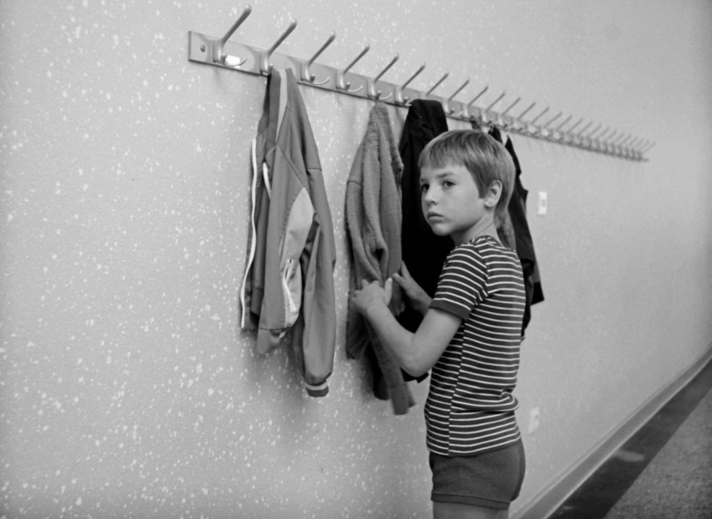 Still from the film Reifezeit (Time of Maturity). A young white boy stands by some hanging coats.