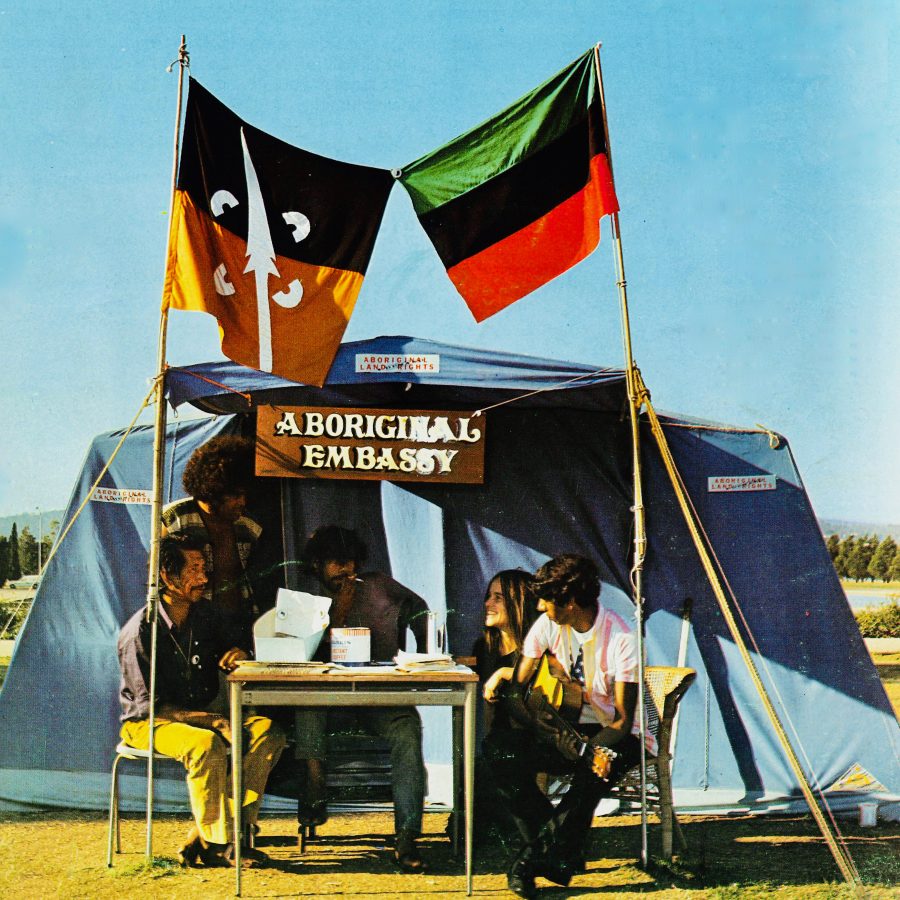 Still from the film Still We Rise. Showing The Aboriginal Tent Embassy  1972. Five Aboriginal people sit in front of a blue tent with flags flying above them.