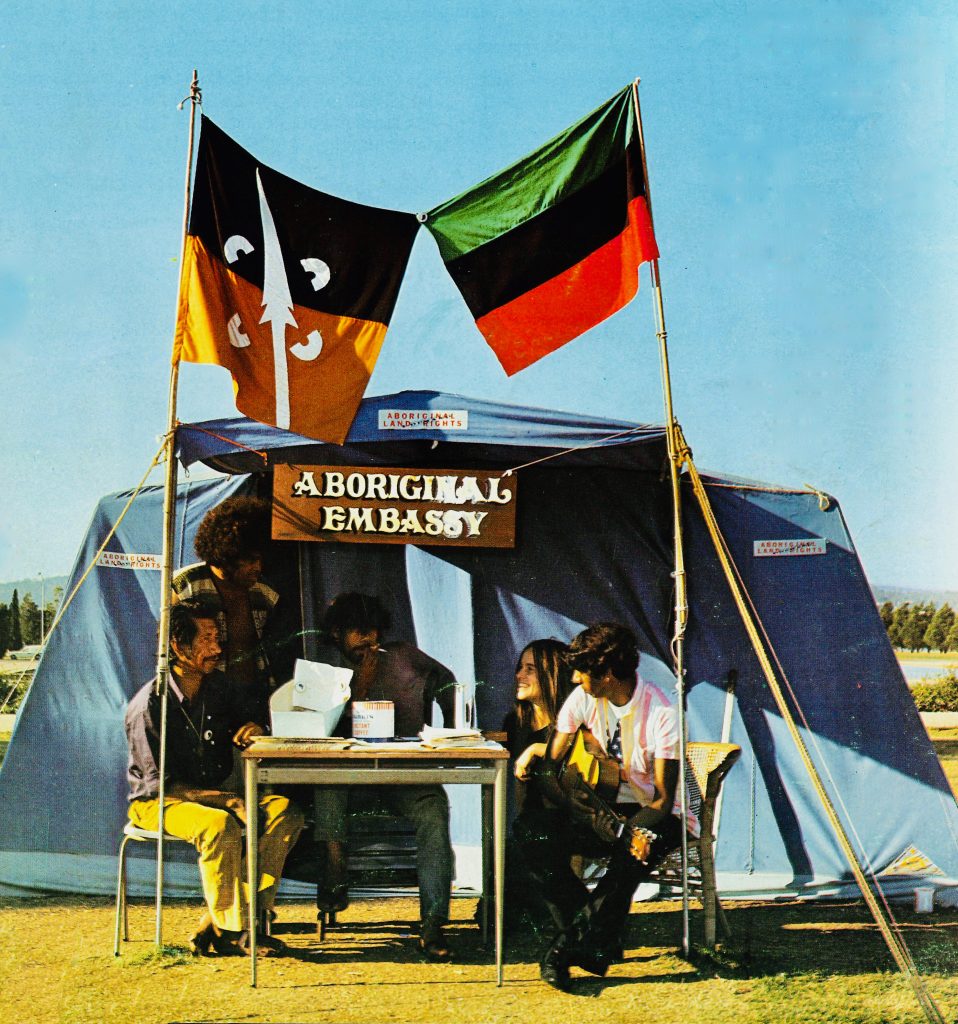 Still from the film Still We Rise. Showing The Aboriginal Tent Embassy  1972. Five Aboriginal people sit in front of a blue tent with flags flying above them.