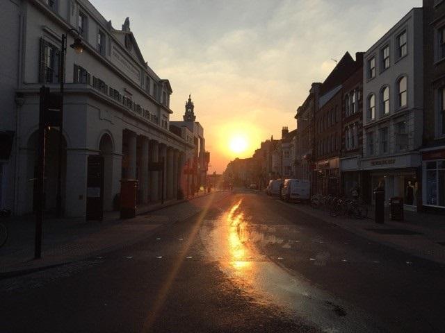 An image taken on a Colchester street at 7am. The image shows the sun rising in the distance which is dimly lighting up the street.