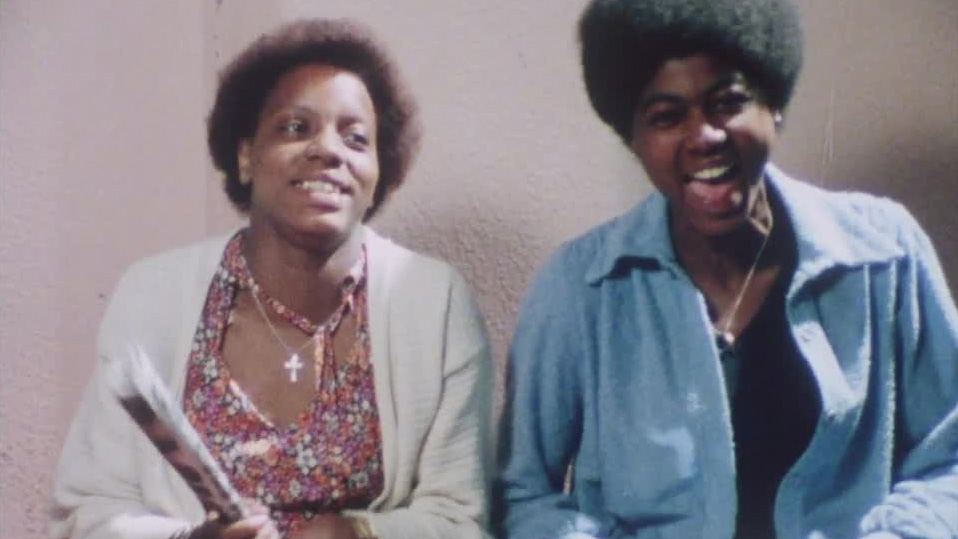 Still from the film Divide and Rule – Never! Showing two black teenagers looking at the camera mid conversation.