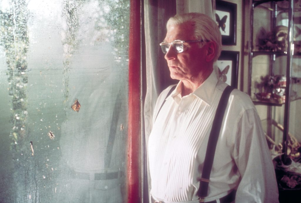 Still from the film Marathon Man. A white man (Laurence Olivier) with white hair stands by a window looking out. There is a butterfly on the window.