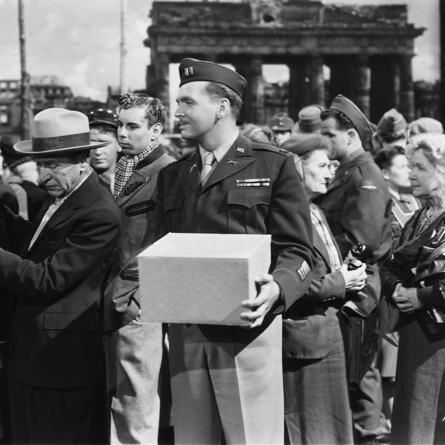 Still from the film A Foreign Affair. A white man in a military uniform stands holding a box. Some people are around him