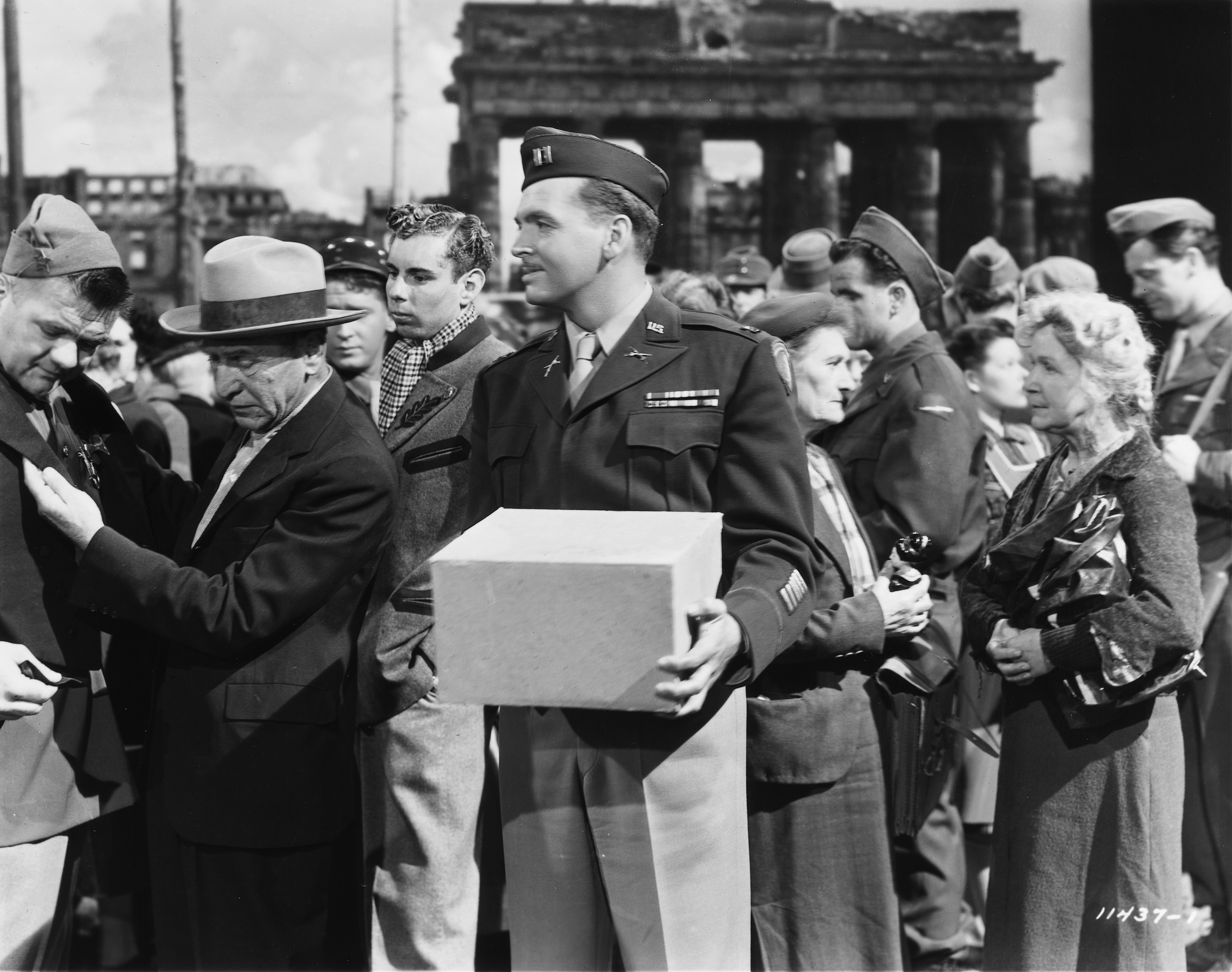 Still from the film A Foreign Affair. A white man in a military uniform stands holding a box. Some people are around him