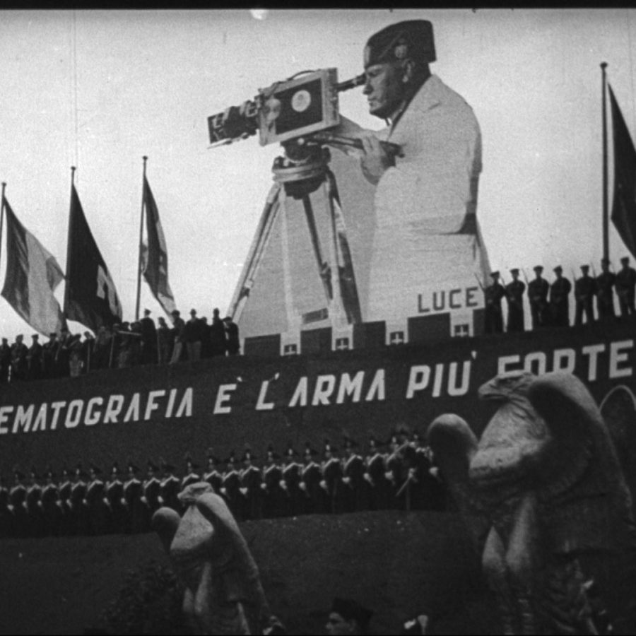 Still from the film The March on Rome. Shows a large representation of Benito Mussolini with a film camera behind a line of fascist soldiers and flags.