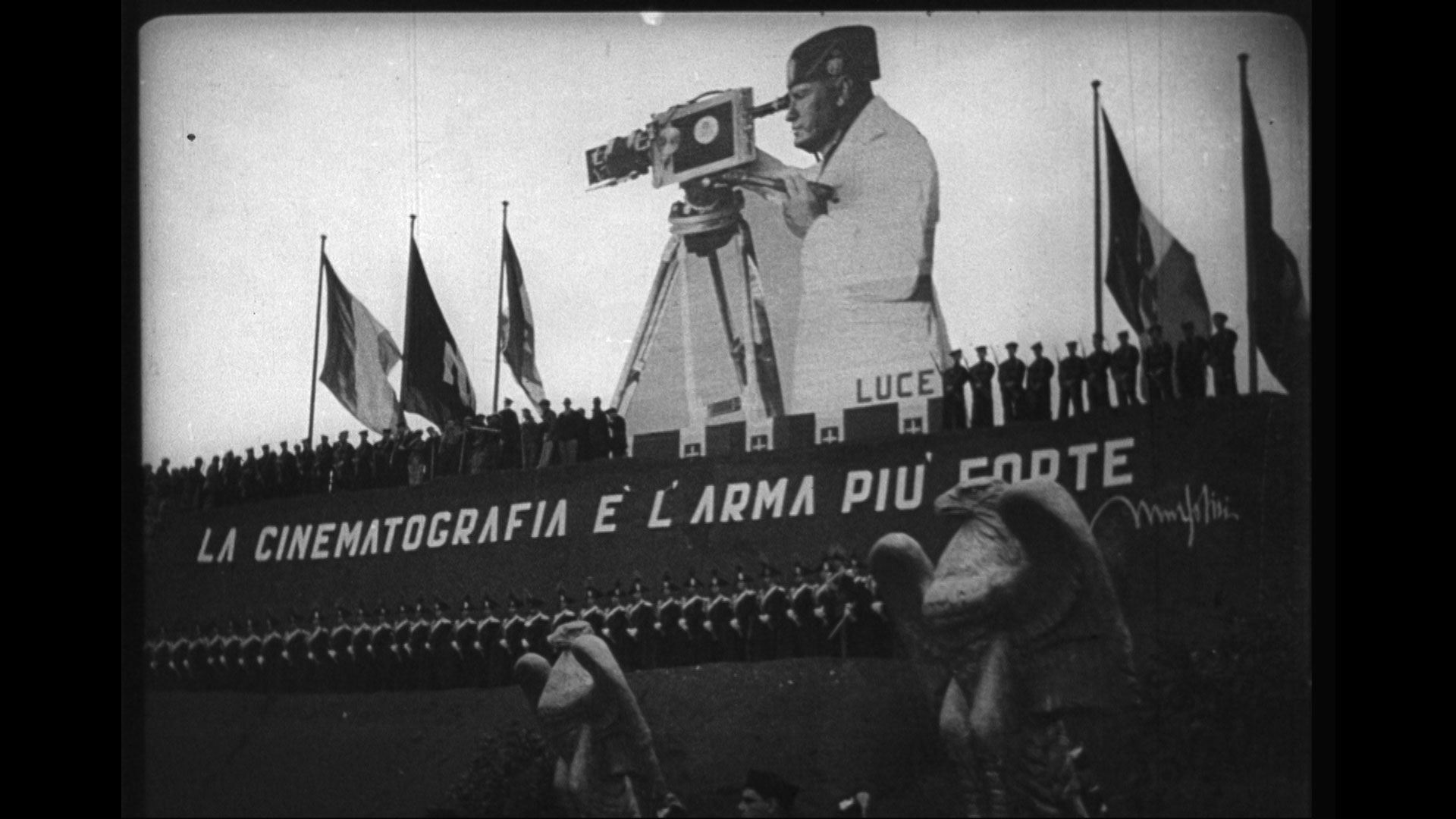 Still from the film The March on Rome. Shows a large representation of Benito Mussolini with a film camera behind a line of fascist soldiers and flags.