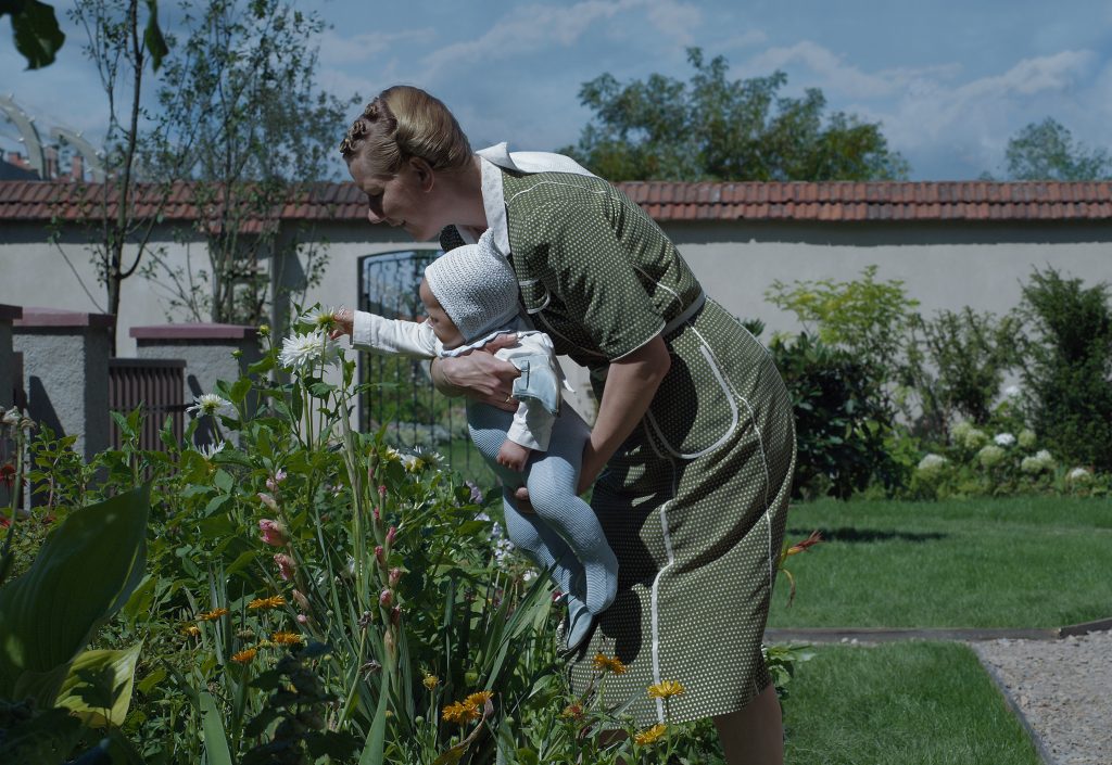 Still from the film The Zone of Interest. A woman a child are picking at a flower in a sunny garden.