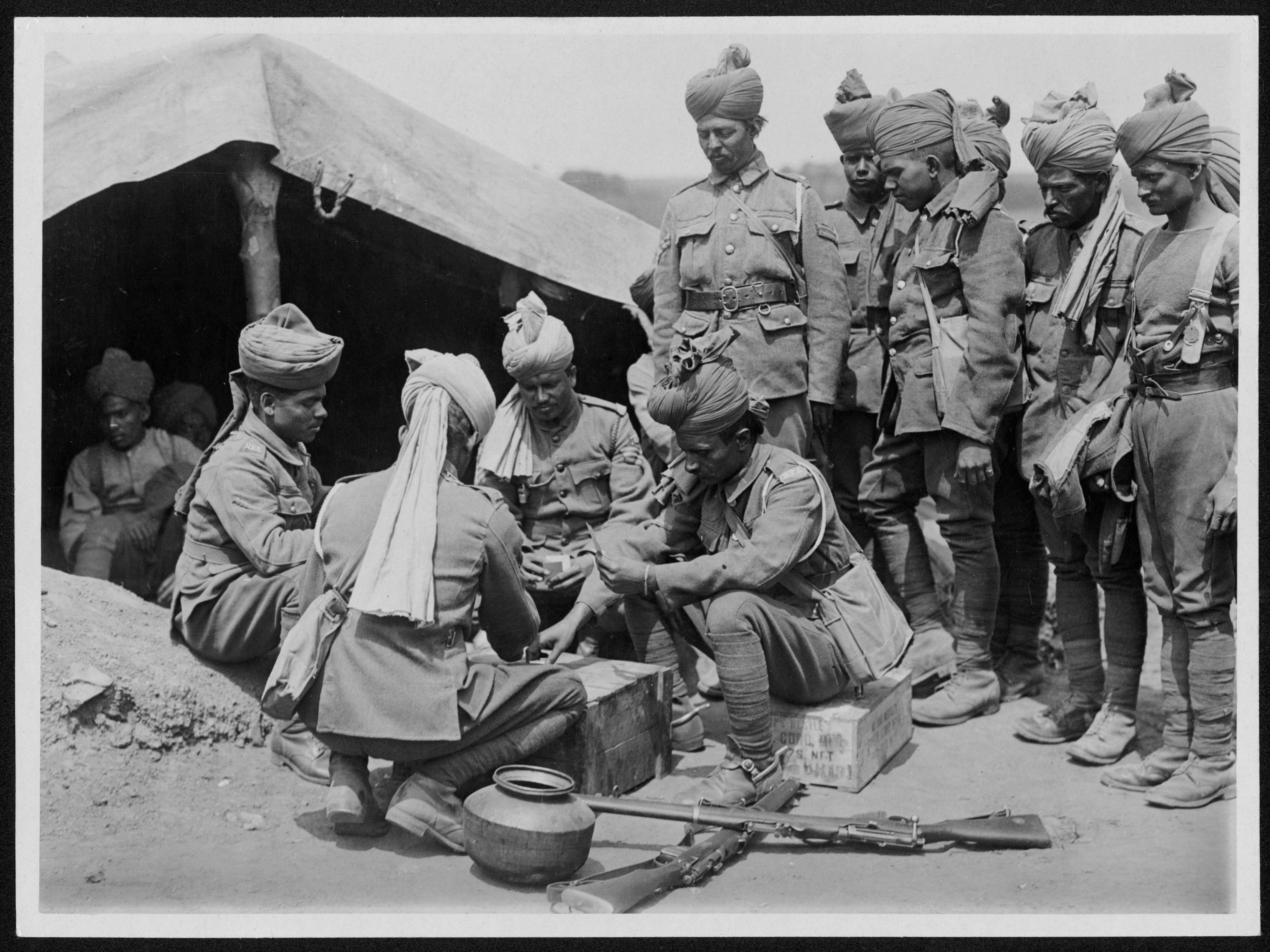 South Asian soldiers playing cards outside a tent. Rifles on the ground.