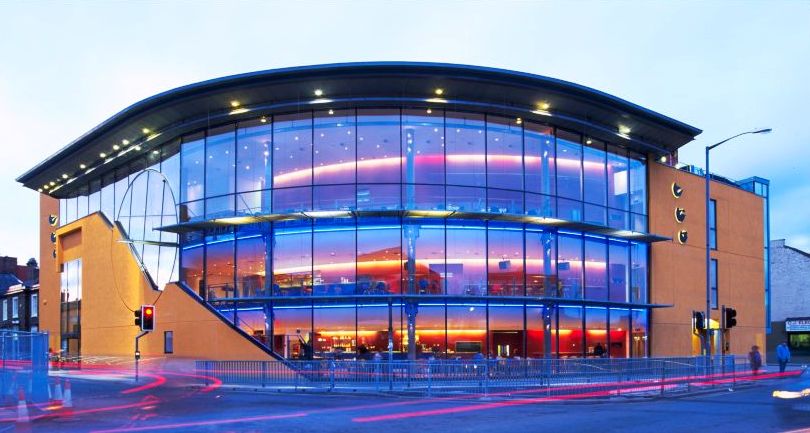 Glass-fronted modern building lit with blue and purple lights at dusk.
