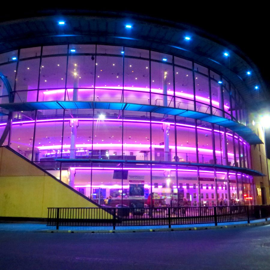 Glass-fronted modern building lit with blue and purple lights at night.