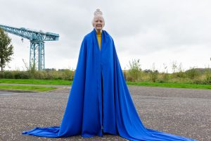 A woman in a long, bright blue cape, with a small crown on her head, stands on a road.
